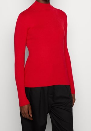 Czerwona, marszczona sweter z długim rękawem i stójką, zestawiona z luźnymi czarnymi spodniami na stojącej osobie na tle jasnego, jednolitego tła.