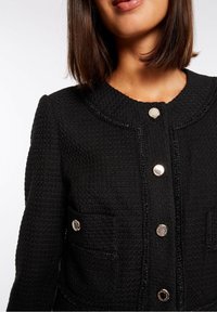 Musta teksturoitu takki pyöreällä kauluksella, viidellä metallisella napilla ja kahdella etutaskulla; saumoissa hienovarainen hohde.