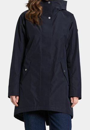 Vrouw draagt een donkerblauwe regenjas met capuchon, vooraan drukknoopsluiting en klepzakken, gecombineerd met donkerblauwe spijkerbroek.