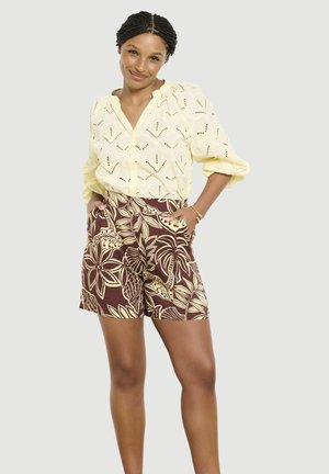 Vrouw met gevlochten haar die een gele eyelet-blouse en bruine korte broek met bloemmotief draagt, handen in de zakken, licht glimlachend.