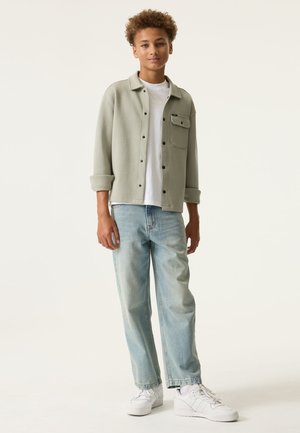 Teenager mit lockigem Haar, trägt eine hellolivgrüne Jacke zum Knöpfen, ein weißes T-Shirt, hellblaue Jeans und weiße Turnschuhe, steht vor einem einfarbigen Hintergrund.