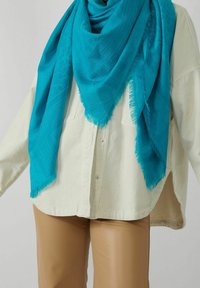 Veste beige boutonnée portée sur un pantalon couleur sable, accompagnée d'une grande écharpe turquoise ornée de motifs subtils et de bords frangés.