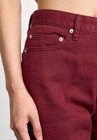 Jeans din denim bordeaux cu o siluetă ajustată, având cinci buzunare, accente din metal argintiu și o textură fină cu nervuri. Prim-plan al zonei taliei.