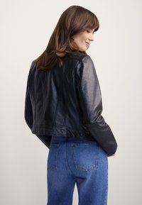 Kobieta o średnio brązowych włosach, ubraną w czarną skórzaną kurtkę i niebieskie dżinsy, spoglądająca przez ramię na jednolitym tle.