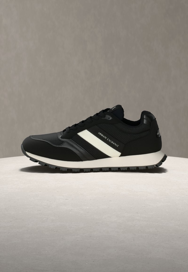 Μαύρο και άσπρο αθλητικό παπούτσι Armani Exchange με μπροστινό δέσιμο με κορδόνια, υφή στη σόλα και λογότυπο στο πλάι και τη φτέρνα, τοποθετημένο σε ουδέτερη επιφάνεια.