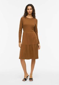 Robe manches longues marron en coton doux avec un haut ajusté et une jupe à volants froncés. Caractérisée par un col rond et une longueur mi-mollet.