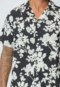 Chemise noire à manches courtes avec un motif floral blanc, dotée d'un col à revers et d'une fermeture à boutons, fabriquée en tissu léger.