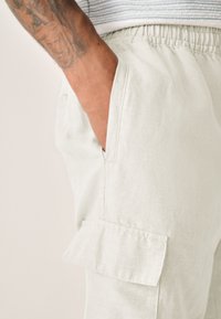 Light grey utility shorts made of textured fabric, featuring side pockets and a cargo pocket with flap detail. Elastic waistband visible.