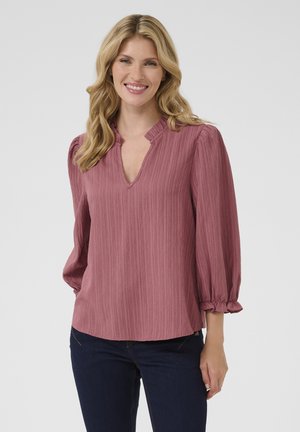 Blusa rosa texturizada con escote en V y mangas abullonadas con fruncidos. Corte casual con puños elásticos y dobladillo recto. Llevada con jeans oscuros.