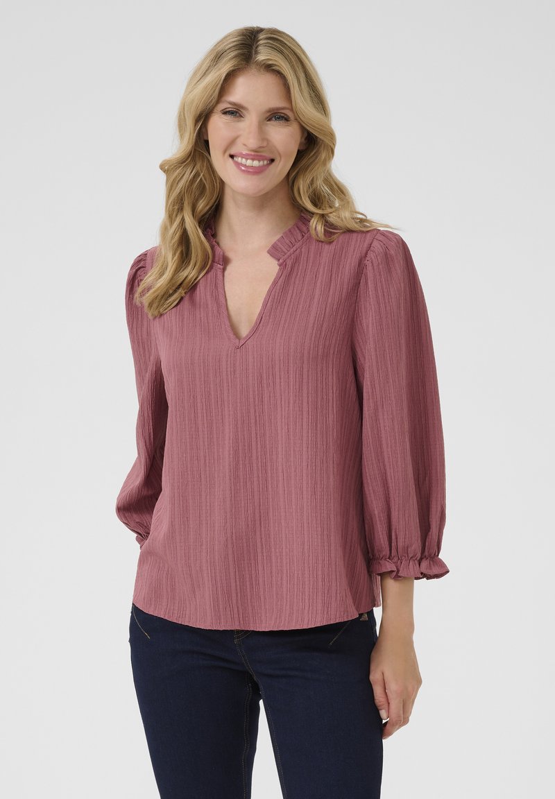 Blusa rosa, strutturata, con scollatura a V e maniche a sbuffo arricciate. Vestibilità casual con polsini elastici e orlo dritto. Indossata con jeans scuri.