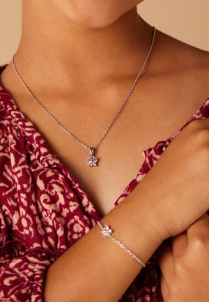 Silberne Halskette und Armbanduhr mit rosafarbenen Edelsteinakzenten, zartem Kettendesign, getragen auf einem gemusterten burgunderfarbenen Stoff.