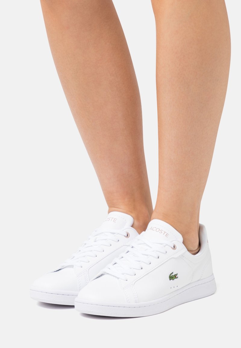Jambes portant des baskets basses blanches Lacoste avec des lacets et un logo crocodile vert sur un fond clair uni.