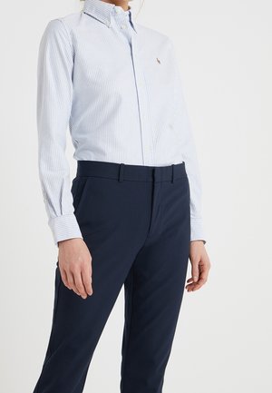 Vrouw draagt een lichtblauw-wit gestreept overhemd met knopen, ingestopt in marineblauwe maatwerk broek, tegen een effen achtergrond.