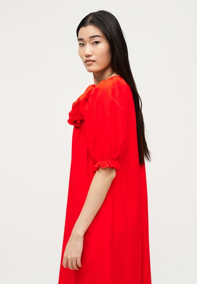 Νεαρή γυναίκα με μακριά μαύρα μαλλιά που φοράει ένα φωτεινό κόκκινο φόρεμα με φουσκωτά μανίκια και βολάν στα μανίκια, κοιτάζοντας πλαγίως σε λευκό φόντο.
