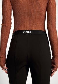 Pantaloni neri con una vita alta che mostra "HUGO" in bianco, abbinati a un top fitted, a maniche lunghe con righe, di colore marrone metallico.