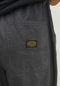 Temno sivi bombažni hlače imajo zadnji žep z črno oznako z napisom "JACK & JONES" in zlatimi poudarki. Tekstura izgleda gladka.