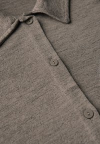 Chemise en tricot gris avec une texture douce, un col et cinq boutons ronds sur le devant. Le tissu présente un motif subtil et varié.