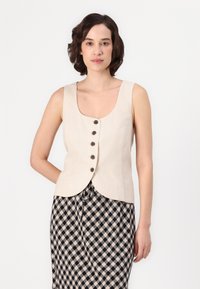 Gilet in lino beige con scollo a barca, cinque bottoni marroni e orlo curvo. Abbinato a una gonna a quadri nera e beige.