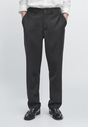 Man wearing dark gray dress pants, black loafers, and a white long-sleeve shirt with hands in pockets, standing on a plain background.