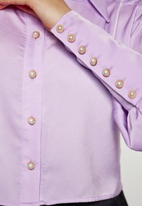 Lavendelbluse i silkeagtig tekstur med perleknapper langs fronten og ærmeenderne, der giver dekorative detaljer.