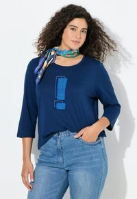 Laivastonsininen pitkähihainen t-paita, jossa etupuolella on teksturoitu sininen huutomerkki. Yhdistetty vaaleansinisiin farkkuihin.