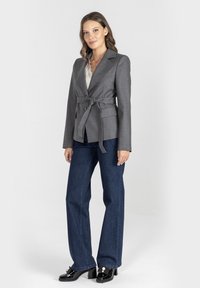 Kobieta w szarym żakiecie z paskiem, białej koronkowej bluzce, ciemnoniebieskich dżinsach i czarnych butach na obcasie z perłowymi detalami, stojąca na białym tle.