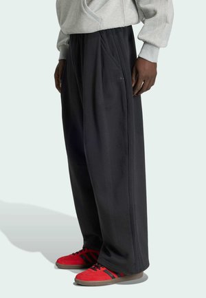 Osoba nosící volné černé kalhoty s logem Adidas, šedou mikinu a červené tenisky Adidas stojící před jednobarevným světlým pozadím.