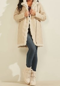 Abrigo beige con patrón texturizado, forro de piel sintética y cierre de cremallera, combinado con vaqueros ajustados azules y botas claras con cordones.