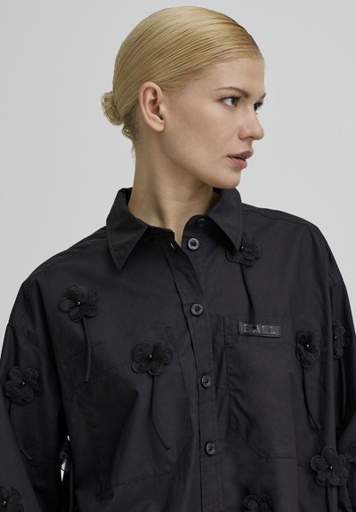 Kvinde med blondt hår samlet i en knold iført en sort skjorte med knapper, dekoreret med sorte stofblomster og en "BALL" mærkat på lommen.
