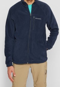 Giacca in pile navy con colletto alto, zip frontale, tasca sul petto con zip e tasche laterali; presenta una texture liscia e cuciture a macchina.