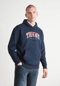 Námornícka modrá mikina vyrobená z hrubej tkaniny, s červenobielym logom "TOMMY JEANS" a kontrastnými červenými šnúrkami. Klokaní vrecko vpredu.