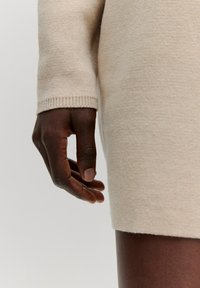 Beige gebreide stoffen jurk met een close-up van een hand die tegen de gestructureerde zoom rust; beschikt over geribbelde mouwdetails voor extra interesse.