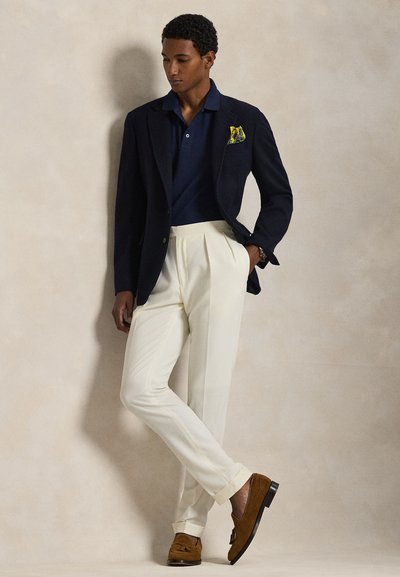 Mies pukeutuneena tummansiniseen bleiseriin, keltaiseen taskuliinaan, tummansiniseen paitaan, valkoisiin laskostettuihin housuihin ja ruskeisiin loafer-kenkiin beigen taustan edessä.