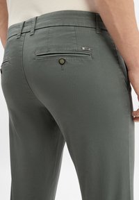 Ciemnoszare spodnie z gładkiego materiału, z dwoma tylnymi kieszeniami zapinanymi na guziki i małą metką marki przy pasie.