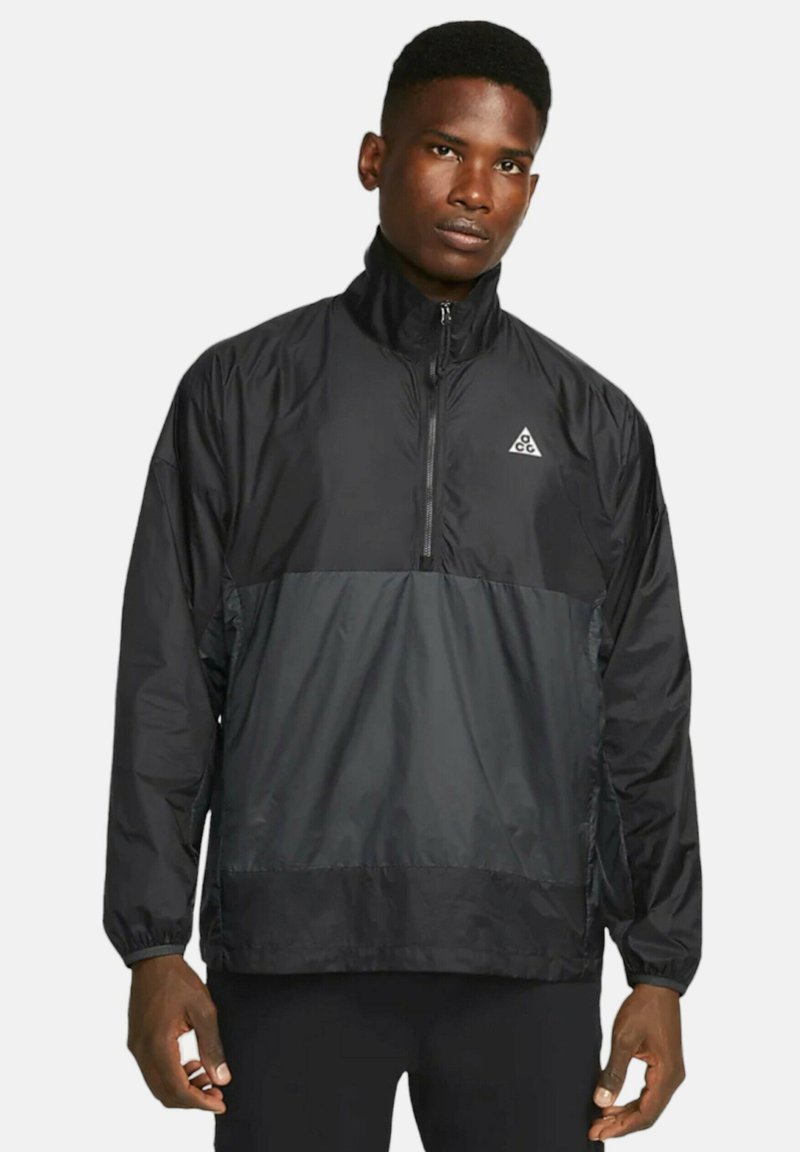 Svart vindjacka med halv dragkedja, med en lättare svart panel över bröstet och ett emblem på vänster sida.