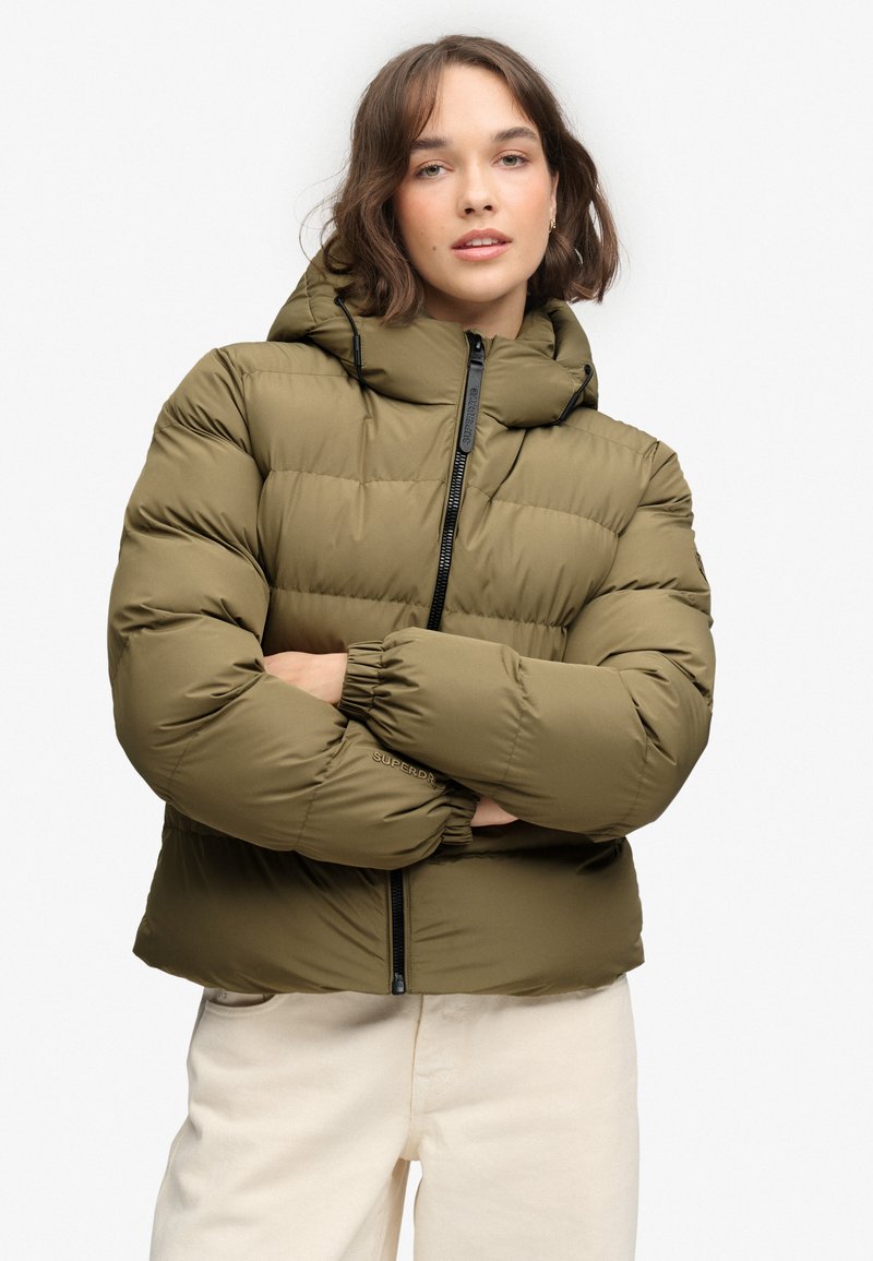 Woman with short brown hair wears an olive green puffer jacket with a hood and cream pants, standing with arms crossed against a plain background.