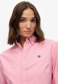 Mujer con cabello castaño hasta los hombros, que lleva una camisa rosa con cuello abotonada y un pequeño logo azul oscuro en el pecho.