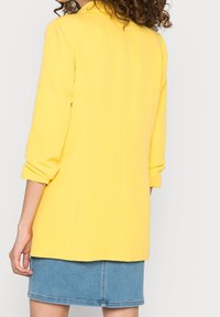 Žena v jasně žlutém saku s vyhrnutými rukávy a krátké modré džínové sukni, zobrazená ze zadní strany na jednobarevném pozadí.