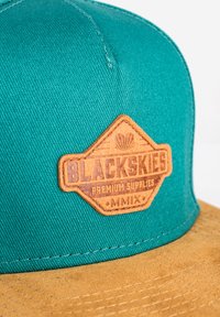 Teal színű anyag sapka barna bőr címkével, amelyen a "BLACKSKIES PREMIUM SUPPLIES MMIX" felirat domborúan, gyémánt alakban található.