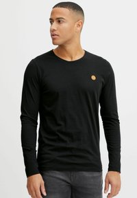 Black long-sleeve t-shirt made of cotton. Features a round neck and a small circular emblem on the chest. Smooth texture and fitted design.