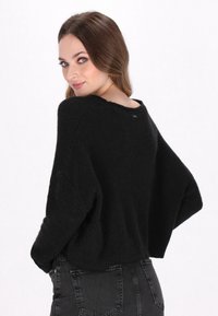 Pull en tricot noir avec une coupe décontractée, des manches larges et un décolleté large, présentant un motif texturé. Associé à un jean gris foncé.