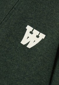 Temnozeleni volneni material z mehko teksturo ima belo, tkano oznako. Oblikovanje vključuje dve prepleteni črki "W".