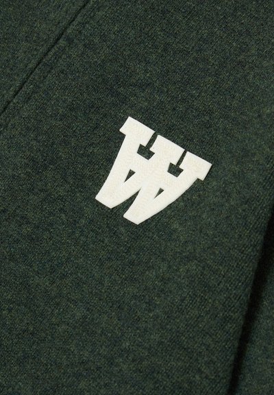 Tmavě zelená vlněná tkanina s měkkou strukturou obsahuje bílou, tkanou nášivku s logem. Design zahrnuje dvě vzájemně propojené písmena "W".