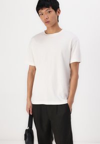 T-shirt en coton blanc à manches courtes avec un col rond, présentant une coupe décontractée. Associé à un pantalon sombre, tenu d'une main par un modèle.