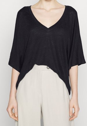 Kobieta ubrana w luźną czarną bluzkę z dekoltem w serek i szerokimi rękawami, lekko wpuszczoną do wysokich, jasno-beżowych spodni.