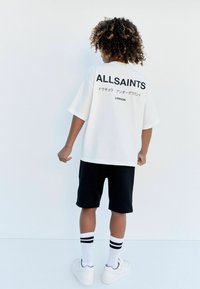 Vit överdimensionerad t-shirt med "ALLSAINTS" logotyp på baksidan, i kombination med svarta shorts, vita sneakers och randiga korta strumpor.