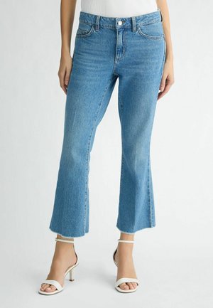 Jeans flare in denim blu chiaro con vita alta, caratterizzati da orlo grezzo e design a cinque tasche, abbinati a tacchi bianchi con cinturini.
