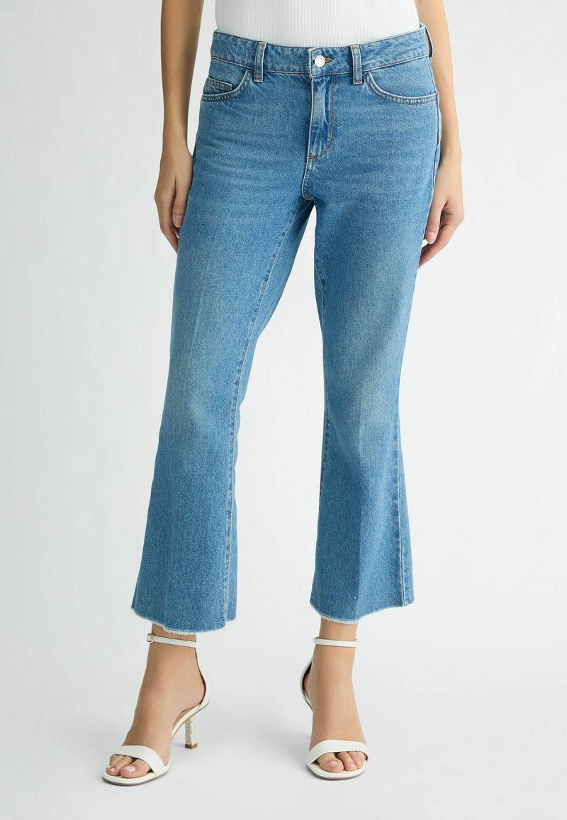Svetlo modri flared jeans z visokim pasom, z raw hemom in petimi žepi, v kombinaciji z belimi strappy petami.