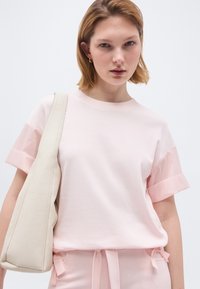 Νέα γυναίκα με κοντά ξανθά μαλλιά που φοράει ανοιχτό ροζ μπλουζάκι και κρατά μια μεγάλη ώχρα λευκή τσάντα ώμου, κοιτάζοντας μπροστά.