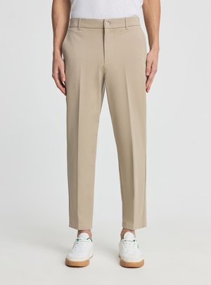 Pantaloni beige a gamba dritta indossati con sneaker bianche e camicia bianca, modello in piedi davanti a uno sfondo chiaro e uniforme.